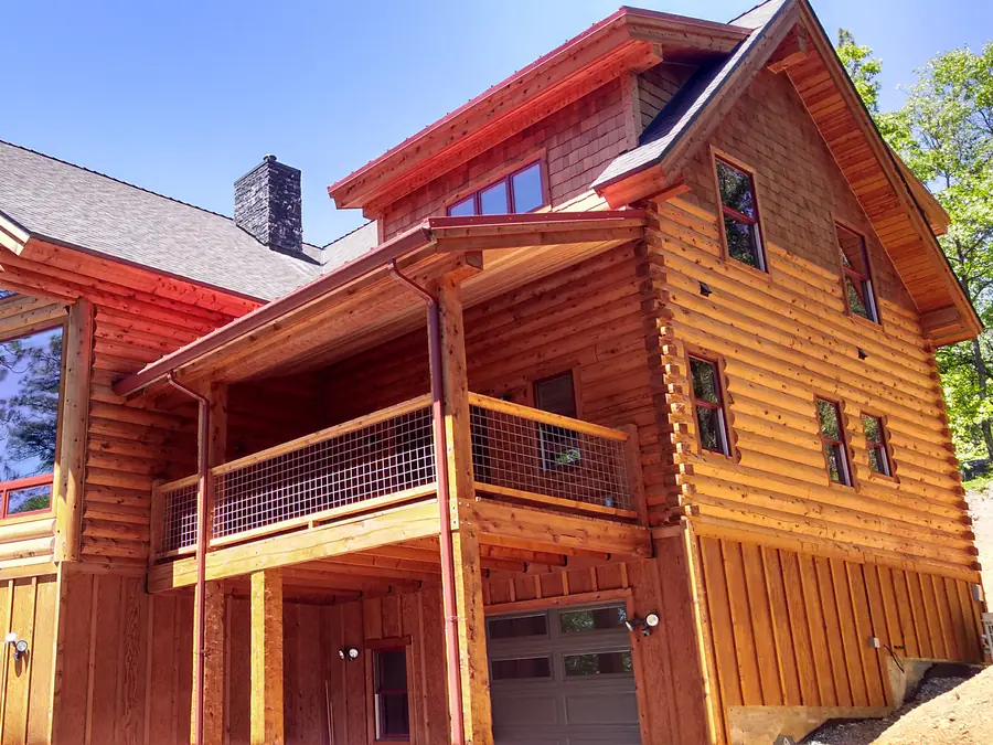 A two-storey D-log home with tall gable windows and a deck, the rounded courses in winter sun.