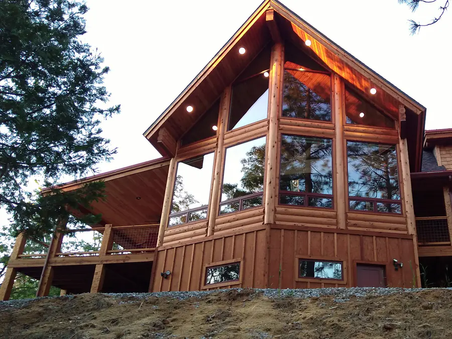 A D-log home with a glazed prow gable and a deck on log posts, at the edge of the woods.