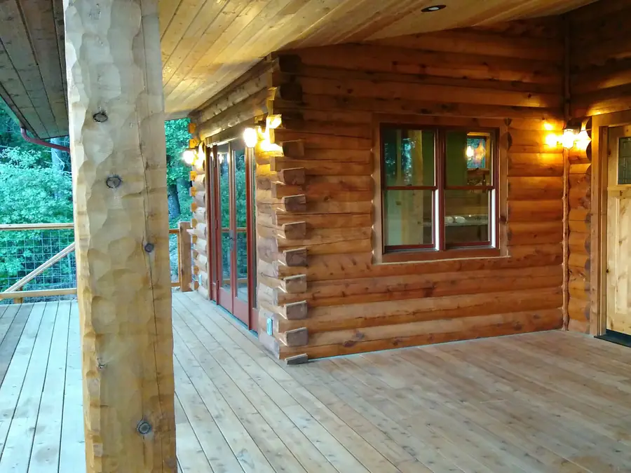 A covered porch along a D-log wall, the rounded courses running past a window and the front door.