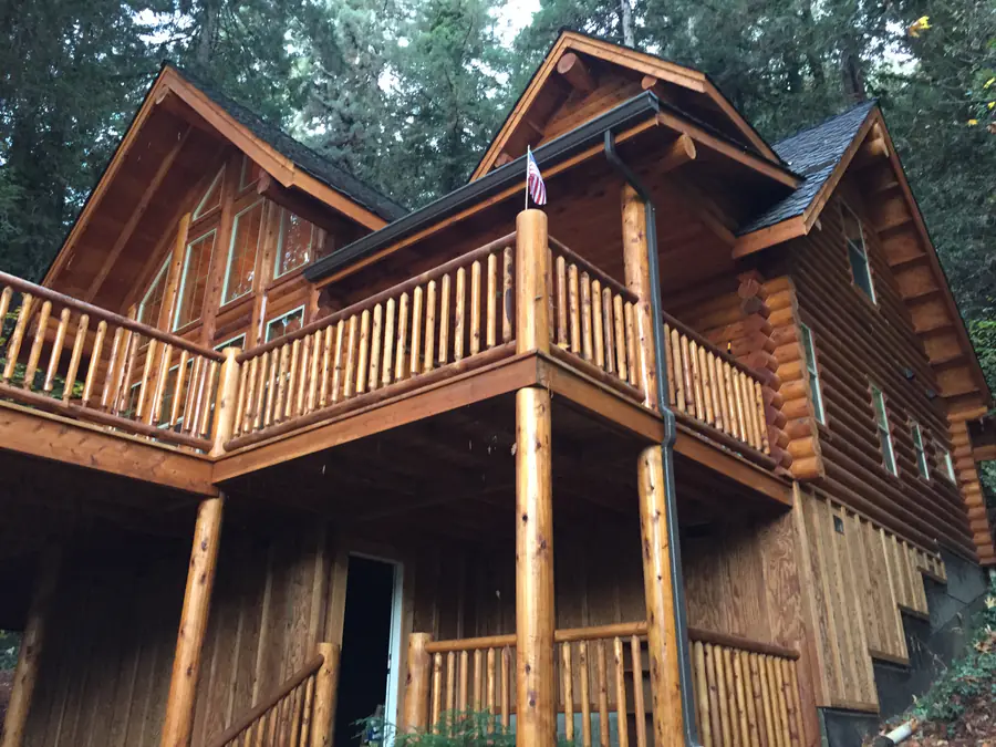 A two-storey round log home with a wraparound deck on log posts, the peeled courses running the full height of the wall under a steep gable.
