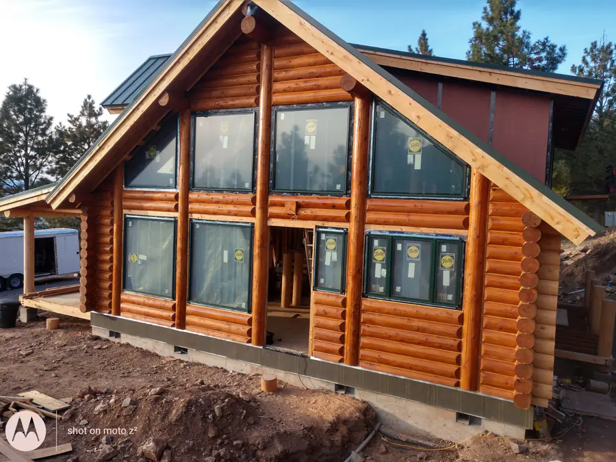 A prow-front round log home with a wall of windows in the gable and saddle-notched log ends stacked at the corners, on a freshly graded pad.
