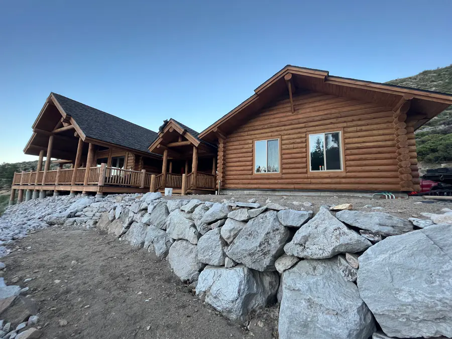 A round log home on a bank of boulders, a covered porch on log posts and the peeled courses lit by low sun.