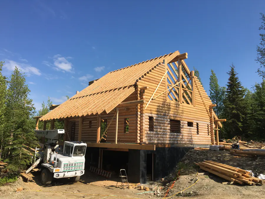 The shell of a round log home with its roof framed, a telehandler at the foundation and log bundles on the ground.