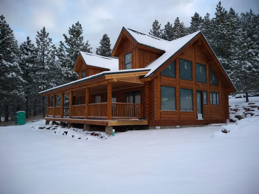 A round log home with a covered porch under fresh snow, the peeled courses running to a prow of windows.