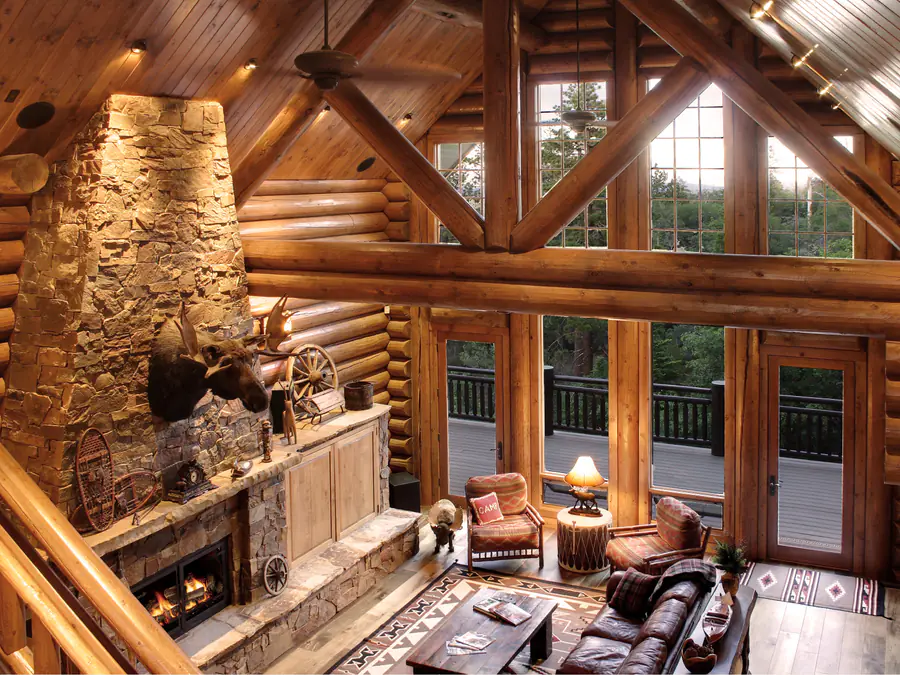 Inside a round log home: a stone chimney rising past the loft railing, log walls and log purlins framing a wall of windows.