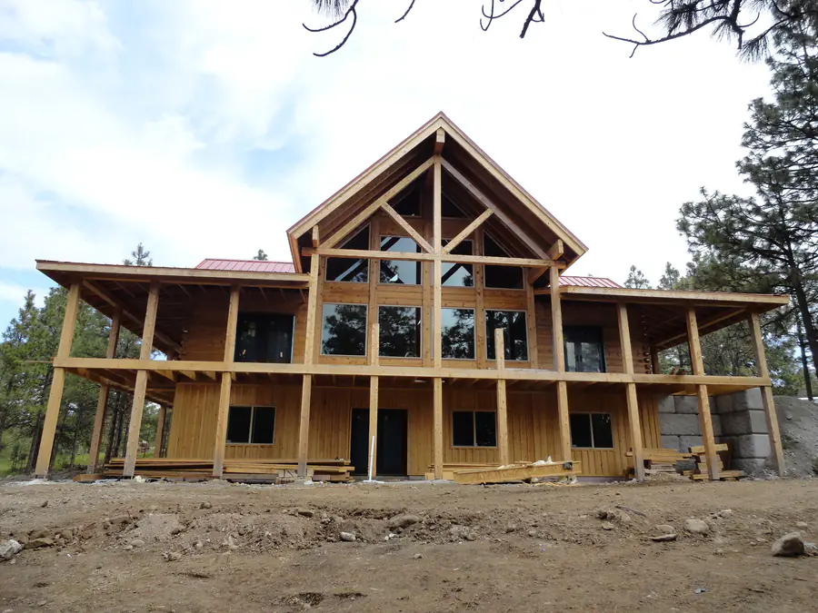A large squared timber home under construction, its glazed gable and two-storey porch framed, on a bare pad.