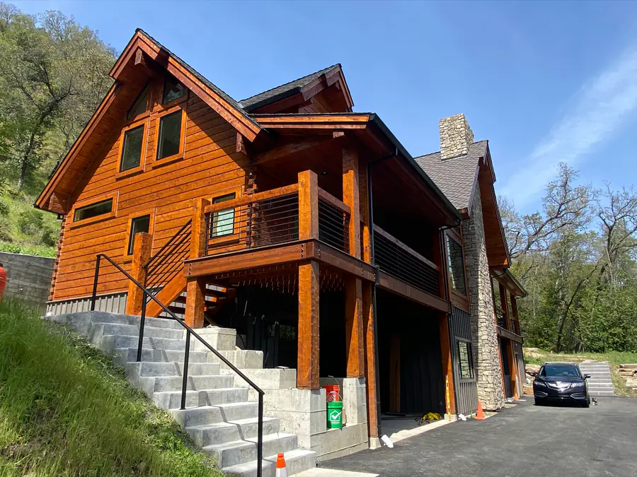 A tall squared timber home with a stone chimney and a deck on log posts, on a hillside above the drive.