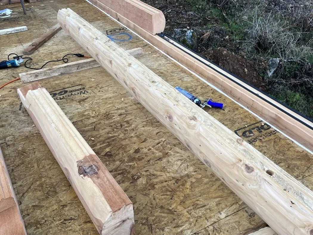 Two long peeled cedar beams lying on a plywood subfloor near the foundation edge, seen from above, with the ground and trees beyond.