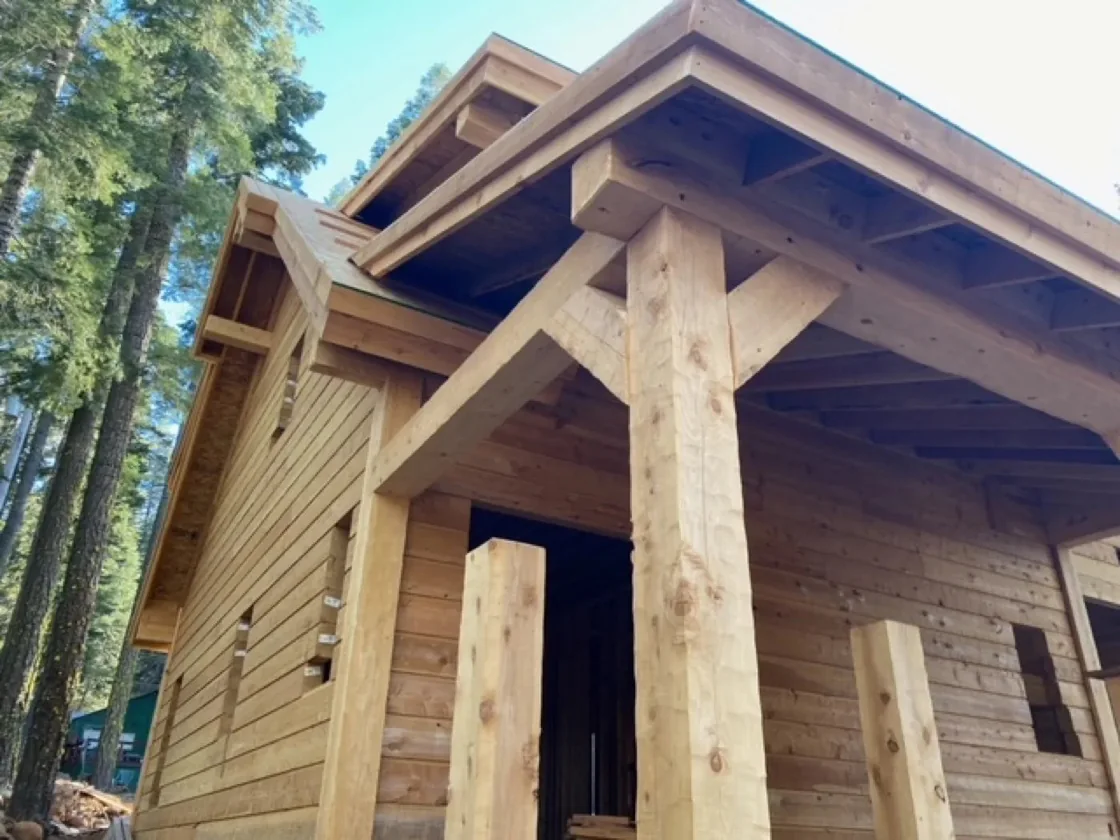 A peeled cedar porch post and a squared beam meeting under a roof overhang, cedar siding behind and pines above.