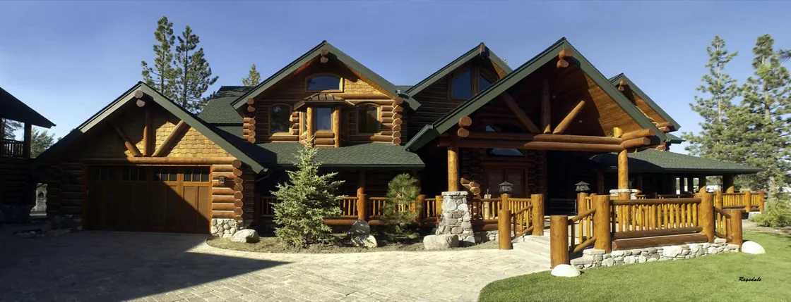 The front of a large finished log home: a gabled entry, a log garage wing, stone piers and a lawn, in full sun.